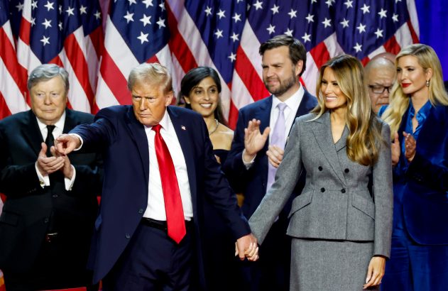 Donald Trump aponta e segura a mão de Melania Trump. Ivanka e outros aplaudem em evento com bandeiras dos EUA.