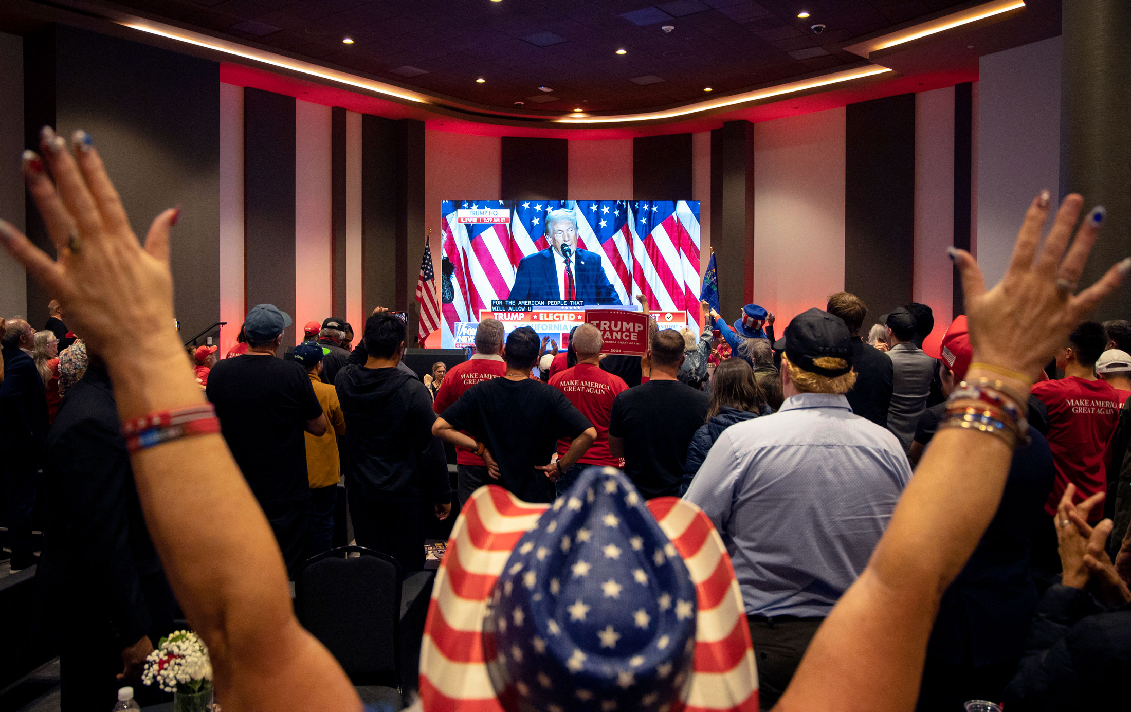 Multidão assiste Trump em telão. Várias pessoas com braços erguidos e vestuário patriótico, incluindo camisas 'MAGA'.