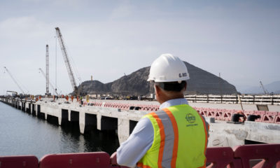 Trabalhador de capacete olha píer em construção com guindastes e montanhas ao fundo.