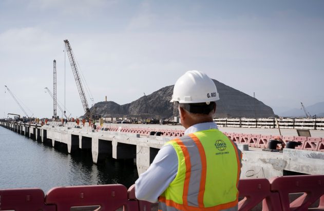 Trabalhador de capacete olha píer em construção com guindastes e montanhas ao fundo.