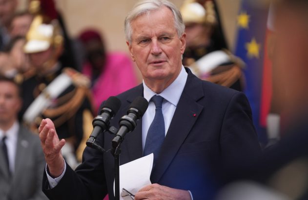 Um homem de terno escuro e gravata azul fala ao microfone, gesticulando. Pessoas e bandeiras ao fundo.