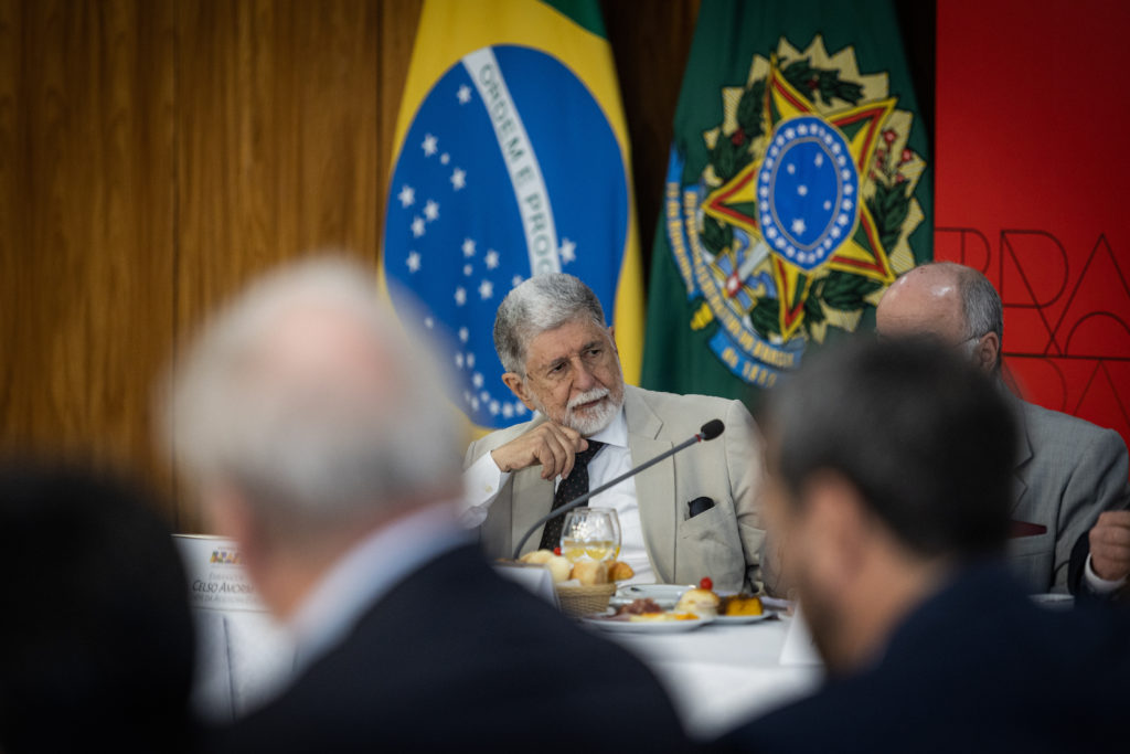 Embaixador Celso Amorim em reunião, olhando atento. Bandeiras do Brasil e brasão nacional ao fundo.