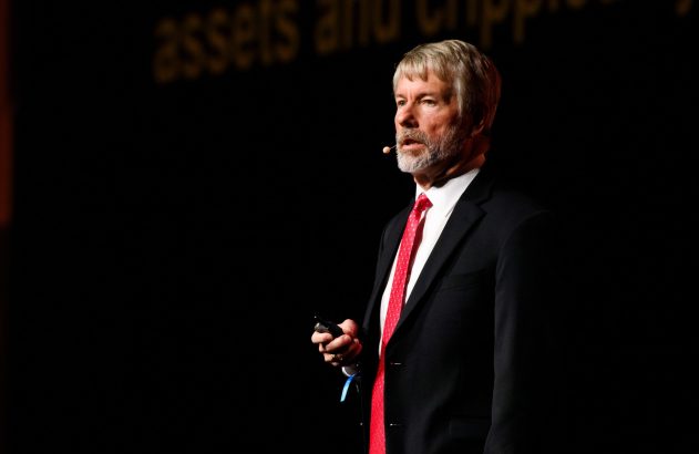 Palestrante em terno e gravata vermelha no palco, segurando um controle, com texto "assets" ao fundo.