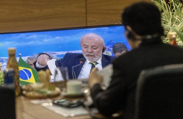 Pessoa assiste o presidente Lula em tela, com bandeira do Brasil na mesa.