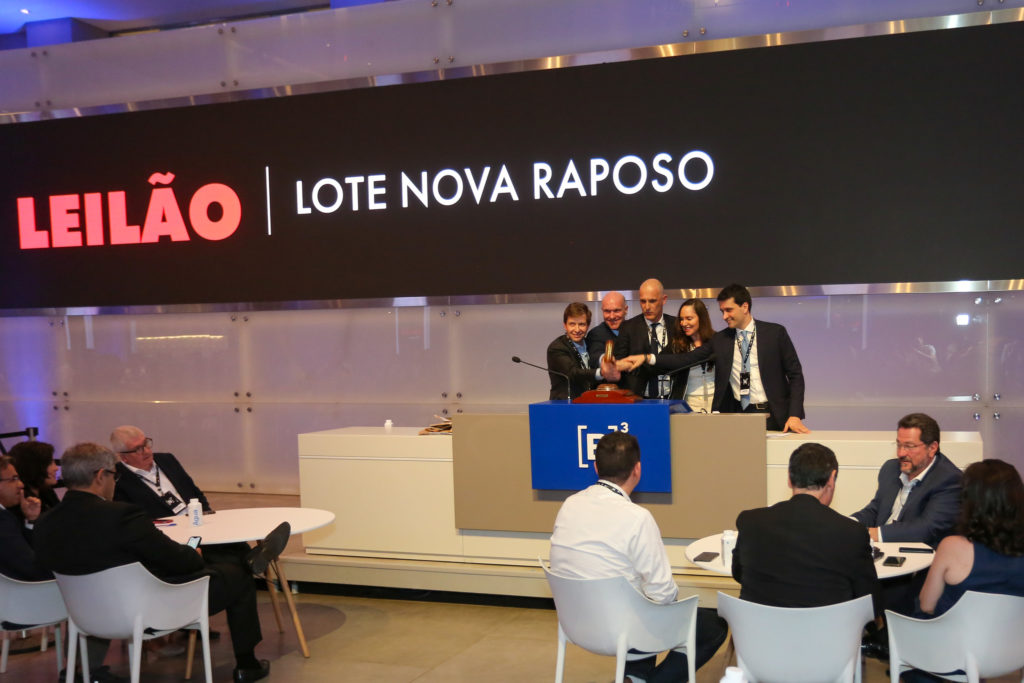 Leilão: Cinco pessoas ativam martelo simbólico no palco. Tela mostra "LEILÃO LOTE NOVA RAPOSO".