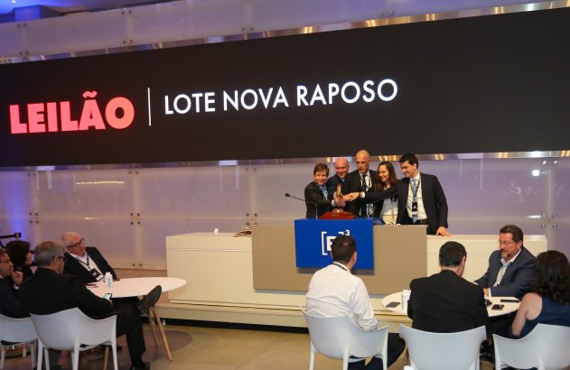 Leilão: Cinco pessoas ativam martelo simbólico no palco. Tela mostra "LEILÃO LOTE NOVA RAPOSO".