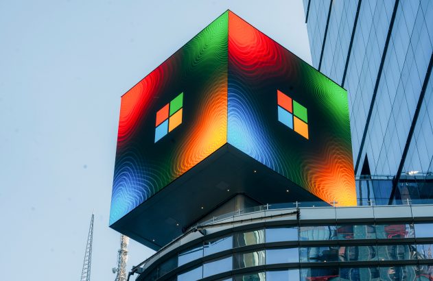 Cubo LED colorido exibindo o logo da Microsoft e cores vibrantes no topo de um prédio moderno.