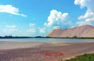Uma paisagem industrial com uma pilha de sal, uma máquina de mineração, uma lagoa azul esverdeada, terreno arenoso e uma fábrica ao fundo, sob um céu parcialmente nublado.