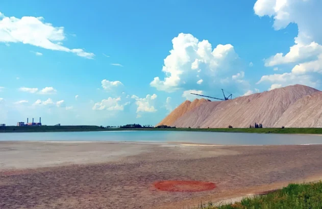 Uma paisagem industrial com uma pilha de sal, uma máquina de mineração, uma lagoa azul esverdeada, terreno arenoso e uma fábrica ao fundo, sob um céu parcialmente nublado.