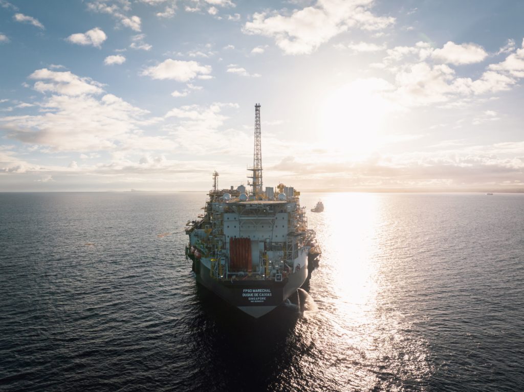 FPSO Marechal Duque de Caxias no mar, rebocador próximo. Sol brilhando baixo no horizonte, céu nublado.