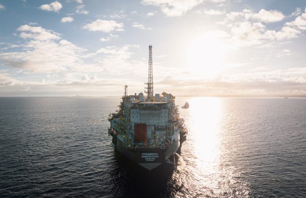 FPSO Marechal Duque de Caxias no mar, rebocador próximo. Sol brilhando baixo no horizonte, céu nublado.