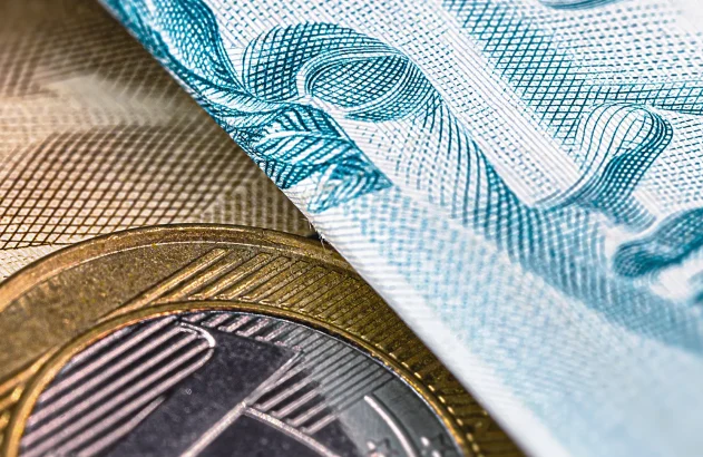 Um close-up de uma cédula azulada e uma moeda bimetálica dourada e prateada, destacando seus detalhes e texturas de segurança.