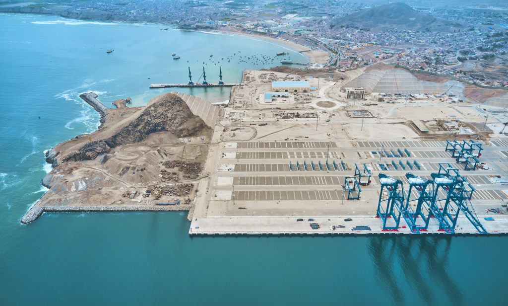Vista aérea de um porto em construção à beira-mar, com guindastes azuis para contêineres e grande pátio.