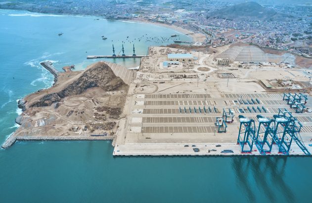 Vista aérea de um porto em construção à beira-mar, com guindastes azuis para contêineres e grande pátio.