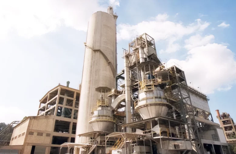 Uma instalação industrial complexa com silos e torres sob um céu azul, cercada por algumas árvores e uma chaminé.