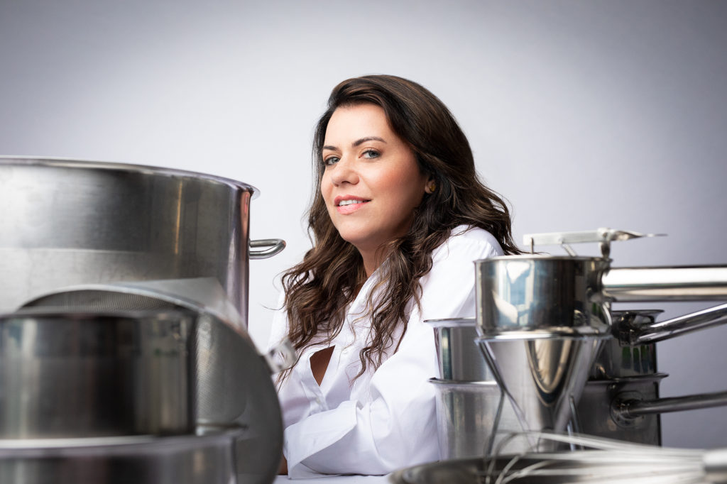 Uma mulher chef sorri entre equipamentos de cozinha industriais de aço inox.