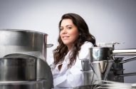Mulher de jaleco branco sorri em ambiente industrial com grandes equipamentos de aço.