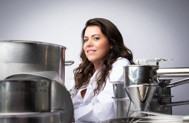 Mulher de jaleco branco sorri em ambiente industrial com grandes equipamentos de aço.