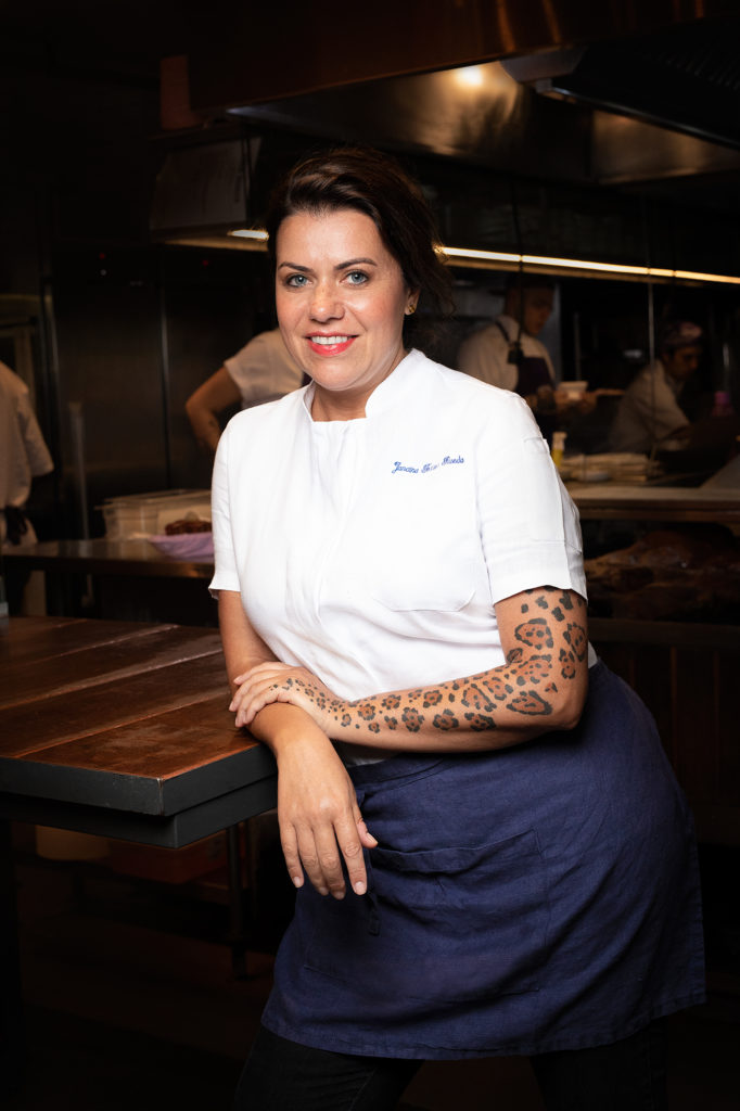 Mulher chef sorri para a câmera, apoiada em uma bancada de cozinha, com tatuagens notáveis nos braços.