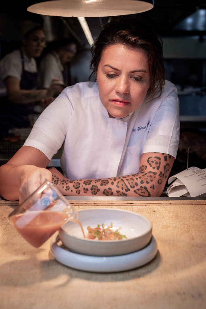 Chef mulher com dólmã branco e braço tatuado com padrão de leopardo, despejando líquido em um prato de comida.