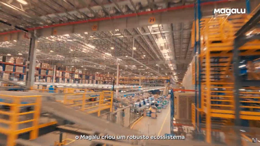 Centro logístico da Magalu: esteiras transportam pacotes, estantes com caixas, operários processam itens.