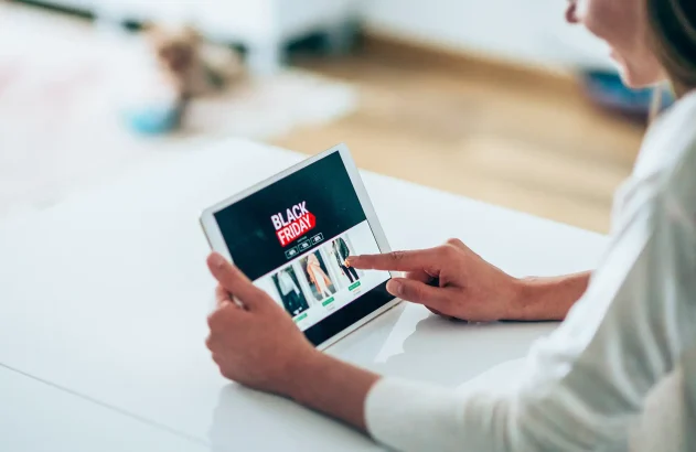 Uma pessoa navegando em uma página de compras online de Black Friday em um tablet sobre uma superfície branca, em um ambiente residencial iluminado.