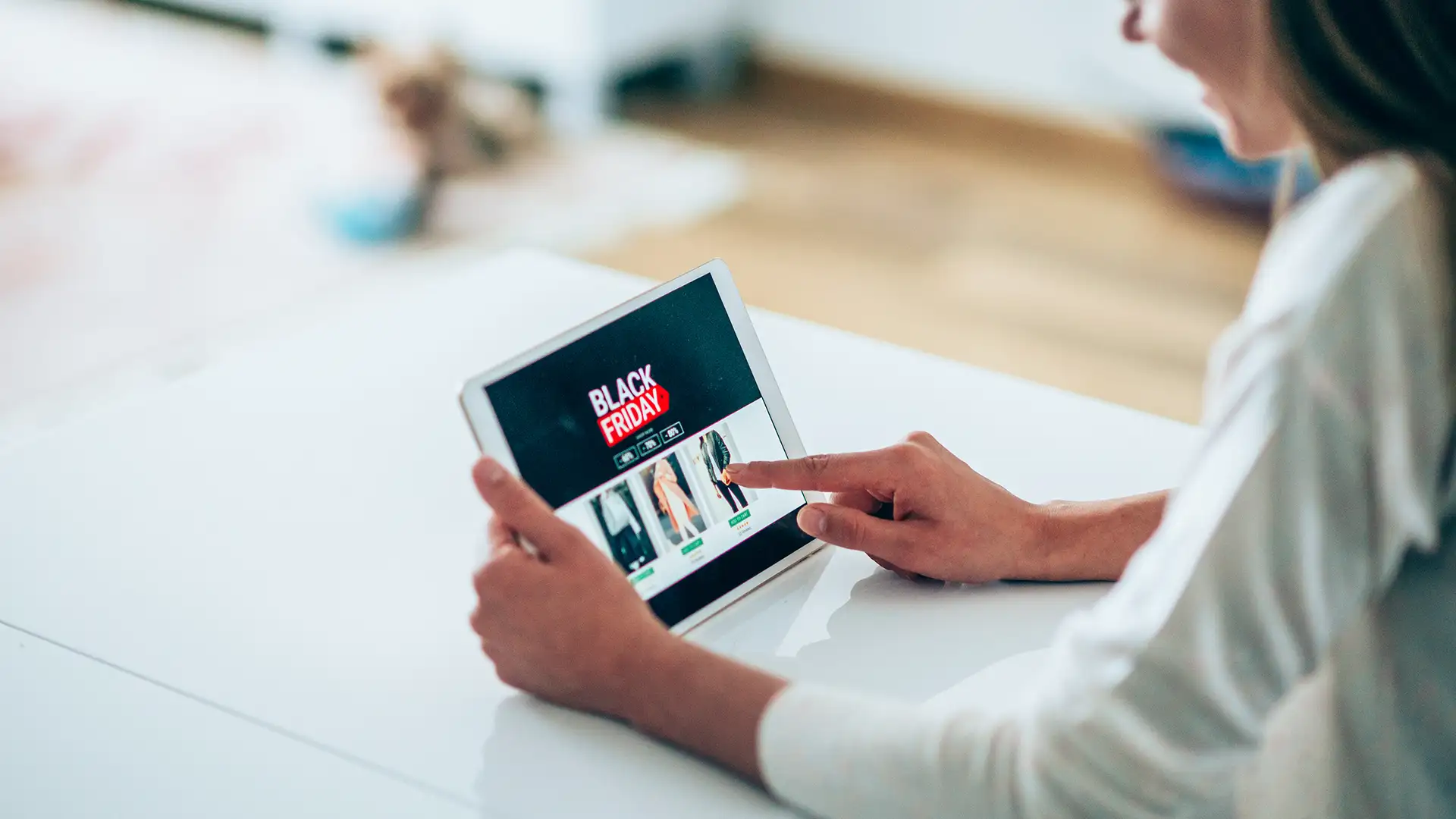 Uma pessoa navegando em uma página de compras online de Black Friday em um tablet sobre uma superfície branca, em um ambiente residencial iluminado.