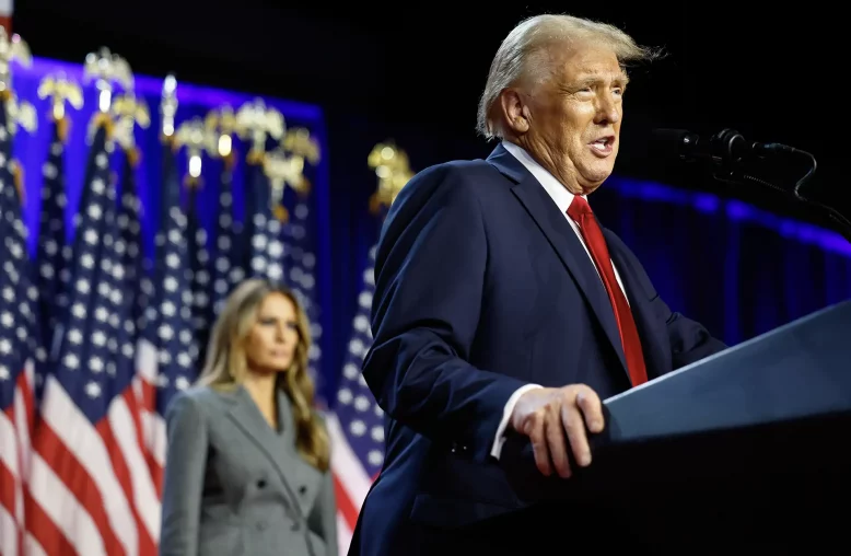 O presidente eleito dos EUA, Donald Trump, um homem em terno azul escuro discursando em um palco com bandeiras americanas ao fundo, observado por sua esposa, Melania Trump, uma mulher de casaco cinza.