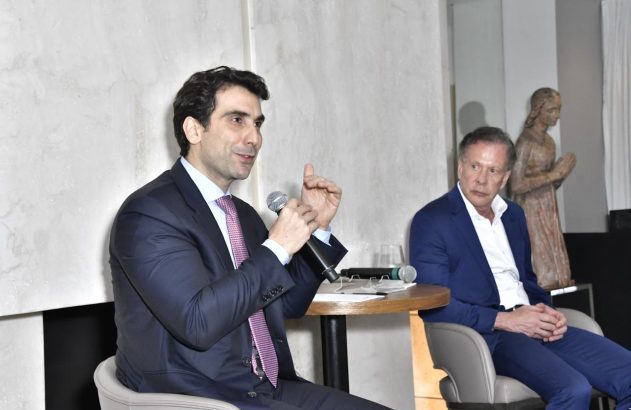 Um homem de terno escuro fala ao microfone, gesticulando, enquanto outro homem o ouve.