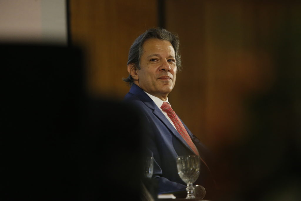 Homem de terno azul e gravata vermelha, cabelos grisalhos, olhando para o lado com leve sorriso.