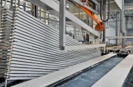 Ambiente industrial com processo de tratamento de metais, estrutura metálica central com lâminas verticais imersas em tanque, maquinário e braços mecânicos ao fundo.
