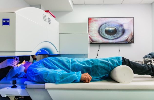 Paciente deitado sob equipamento oftalmológico Zeiss. Um profissional opera o olho, enquanto tela exibe imagem ampliada.