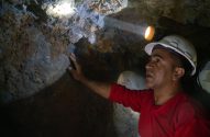 Homem de capacete com lanterna ilumina e toca parede de rocha em túnel escuro de mina, olhando para cima.