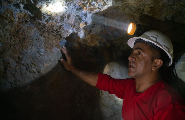 Homem de capacete com lanterna ilumina e toca parede de rocha em túnel escuro de mina, olhando para cima.
