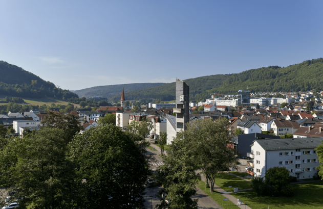 Vista panorâmica de uma cidade em vale verdejante, com casas e torres de igreja e relógio sob céu azul.