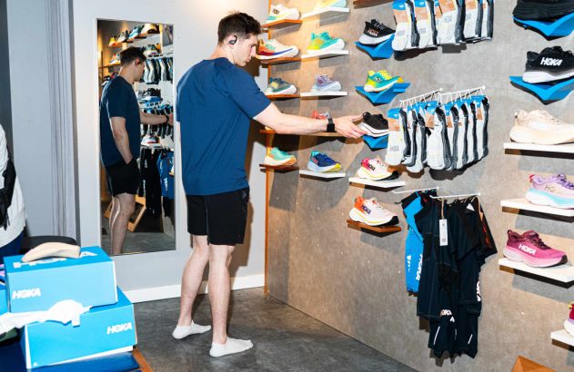 Homem em loja Hoka, tocando em tênis na prateleira, com reflexo no espelho.