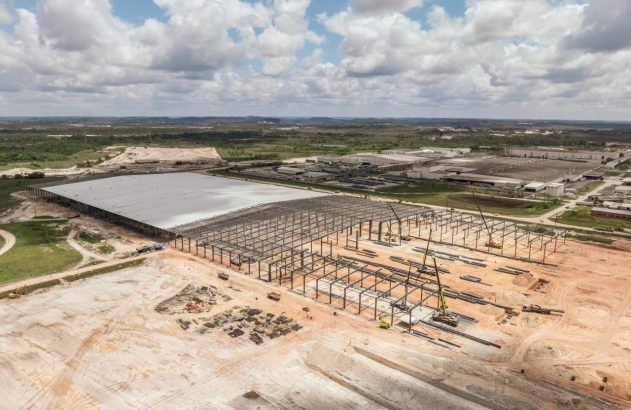 Grande galpão industrial em construção com estrutura de aço e guindastes. Terreno amplo e fábricas ao redor.
