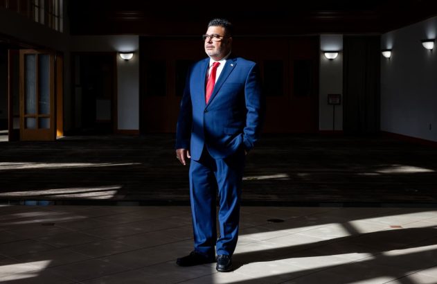 Homem de terno azul e gravata vermelha em pé, mão no bolso, em salão amplo com luz solar no chão.