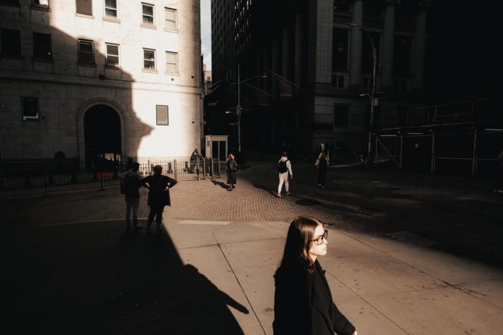 Mulher de óculos no primeiro plano andando em rua urbana com luz e sombra intensas. Pessoas e prédios ao fundo.