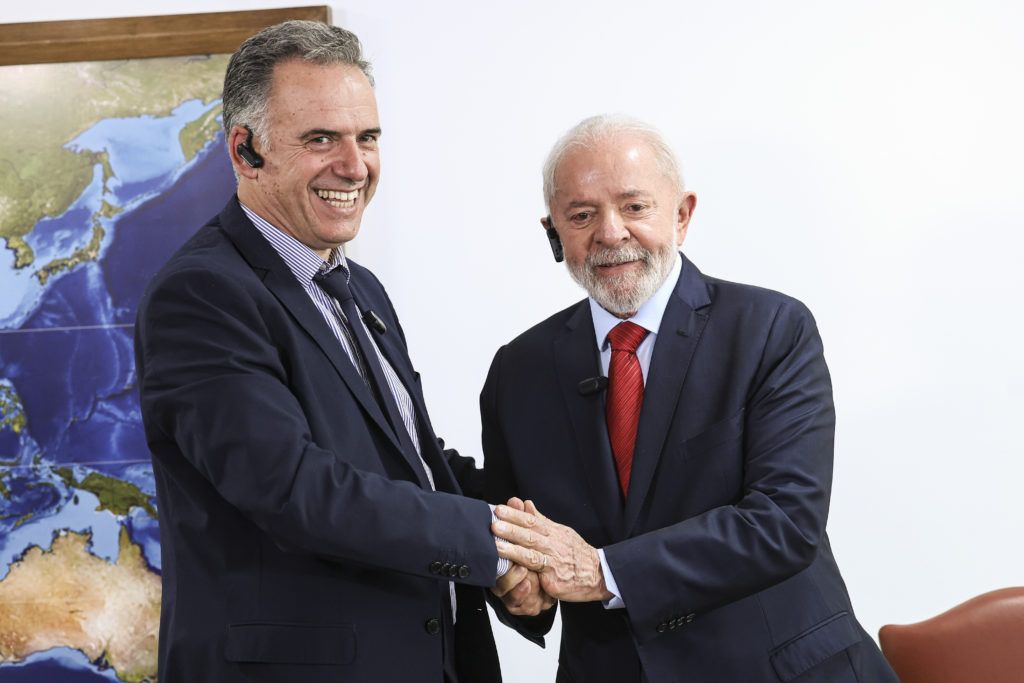 Dois homens sorridentes, de terno e fones de ouvido, apertam as mãos. Há um mapa ao fundo.