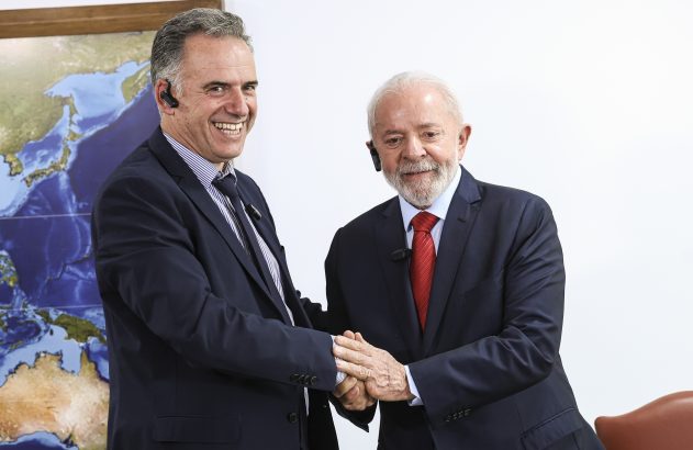 Dois homens sorridentes, de terno e fones de ouvido, apertam as mãos. Há um mapa ao fundo.