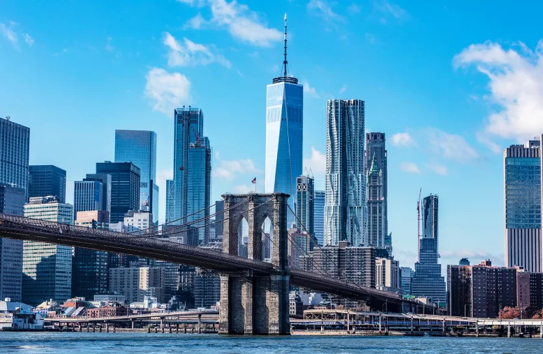 A imagem retrata o skyline de Nova York com a Ponte do Brooklyn em primeiro plano e Manhattan ao fundo, destacando o One World Trade Center contra um céu azul com nuvens esparsas.