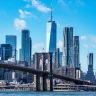 A imagem retrata o skyline de Nova York com a Ponte do Brooklyn em primeiro plano e Manhattan ao fundo, destacando o One World Trade Center contra um céu azul com nuvens esparsas.