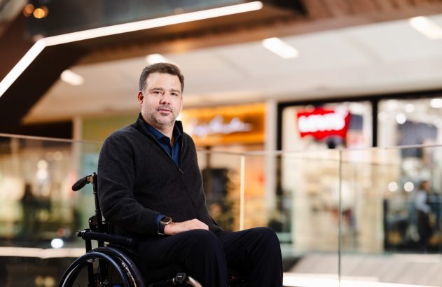 Homem em cadeira de rodas sentado em ambiente de shopping, olhando para a frente.