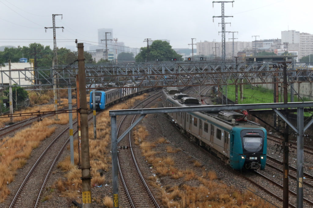 Dois trens em movimento na ferrovia. Um se afasta à esquerda, outro se aproxima à direita. Cenário urbano nublado.