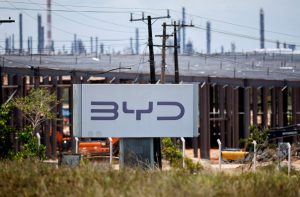 Placa da BYD em canteiro de obras de fábrica, com chaminés industriais e postes ao fundo.