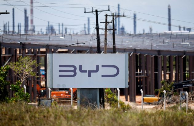 Placa da BYD em canteiro de obras de fábrica, com chaminés industriais e postes ao fundo.