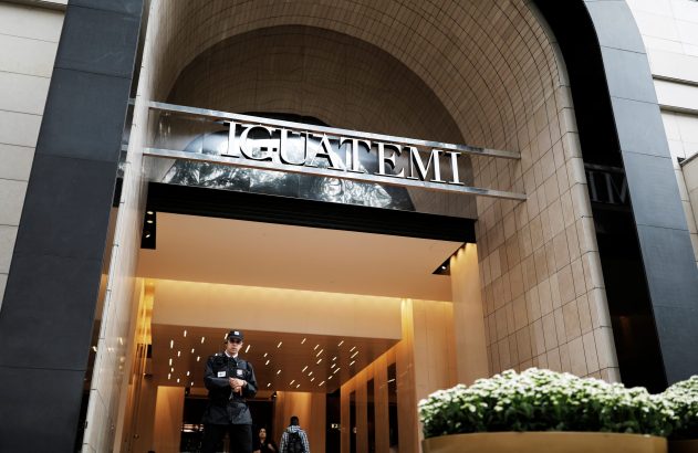 Entrada de edifício moderno com grande arco, painéis pretos e estrutura prateada suspensa. Pessoa parada na frente, outras entram.