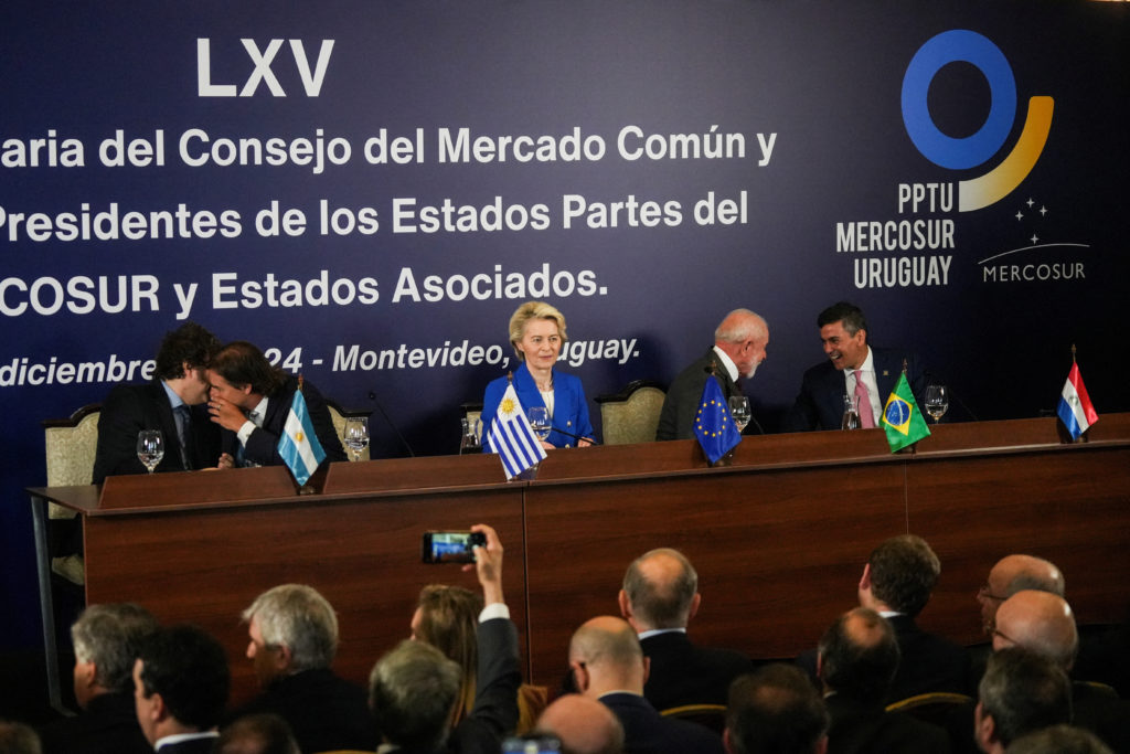 Líderes de países e da UE em cúpula do Mercosul. Ursula von der Leyen e Lula em mesa, com bandeiras e público.