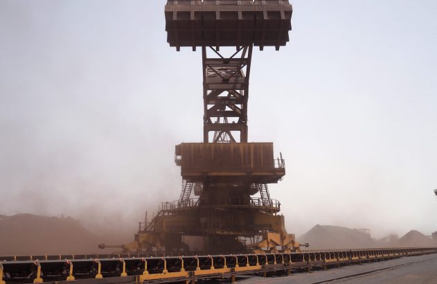 Máquina empilhadeira-recuperadora industrial manuseando minério, levantando poeira, com esteiras.